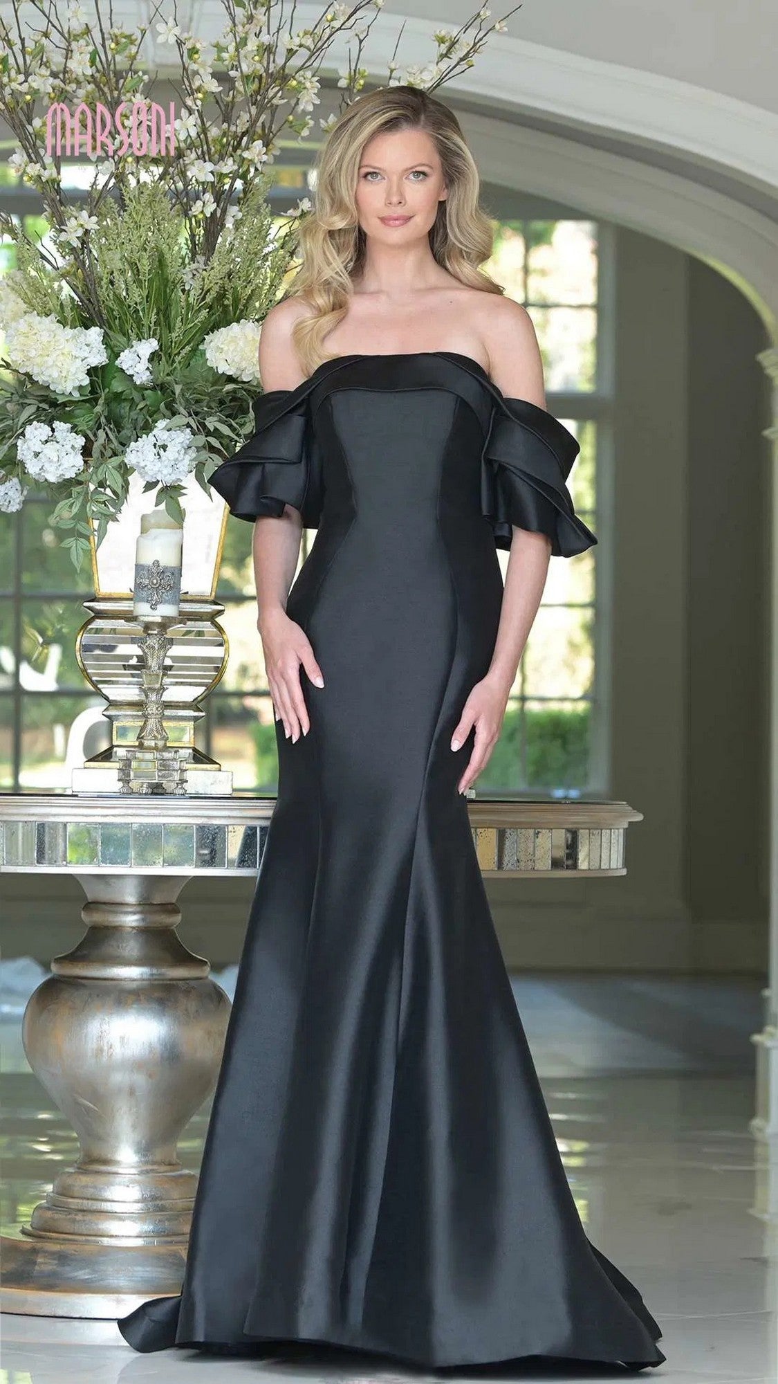 A woman stands indoors wearing the Marsoni MV1290 long black formal dress with off-the-shoulder ruffled sleeves. A large floral arrangement appears behind her.